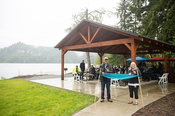 A woman and man perform a ribbon cutting ceremony with people gathered around picnic tables under an open air shelter by Lake Shannon in rainy weather.